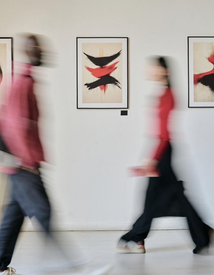 Sonder Two blurred figures in casual attire walk past abstract art pieces displayed on a white wall in a gallery setting.